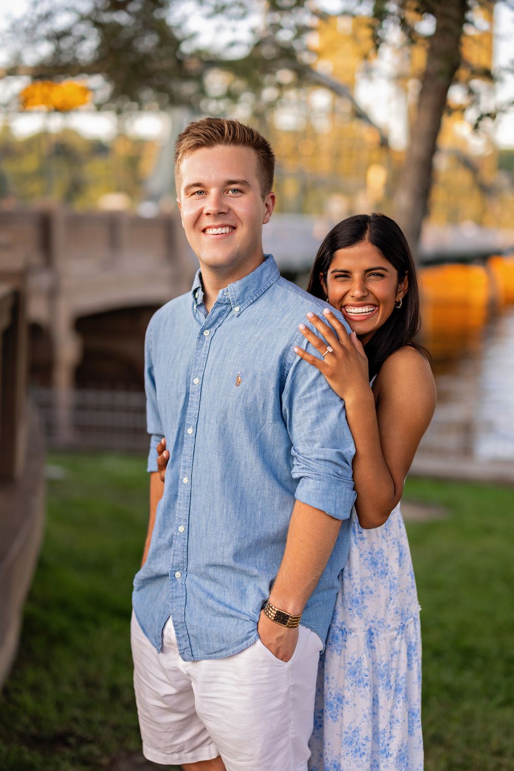 Joyful couple posing back-to-back outdoors near a bridge, woman in floral dress laughing, man in blue shirt.