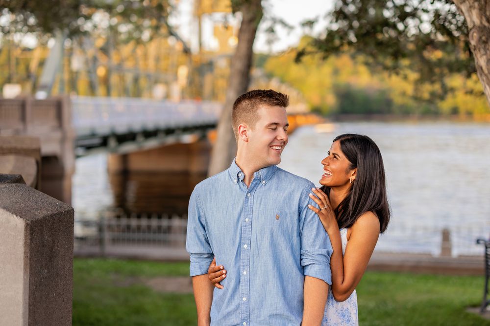 Couple sharing a tender moment outdoors near a waterfront bridge, woman in white dress touching mans shoulder.