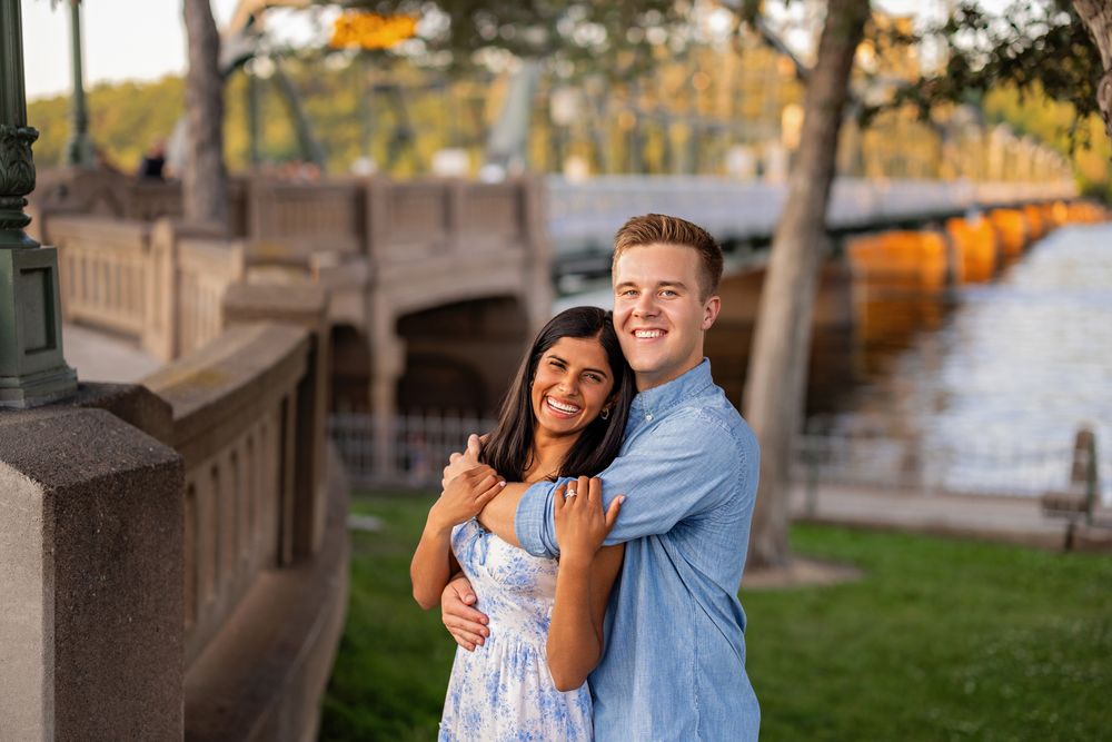 Happy couple hugging and smiling near a scenic waterfront bridge surrounded by lush greenery.