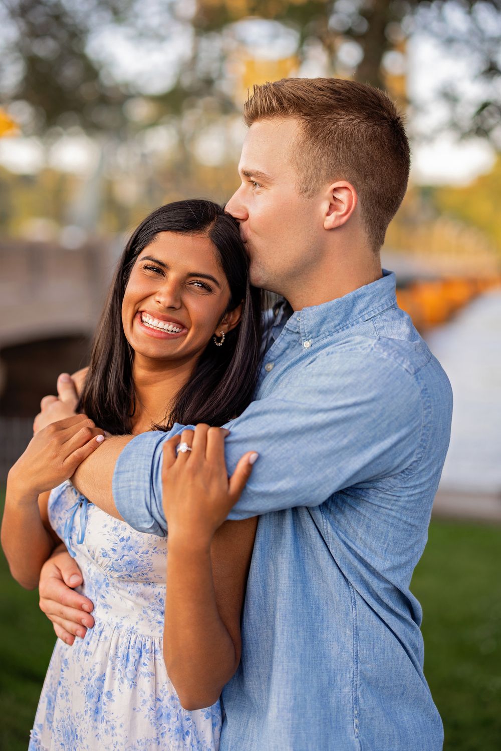 Man in blue shirt kissing womans cheek while embracing her outdoors near a bridge.