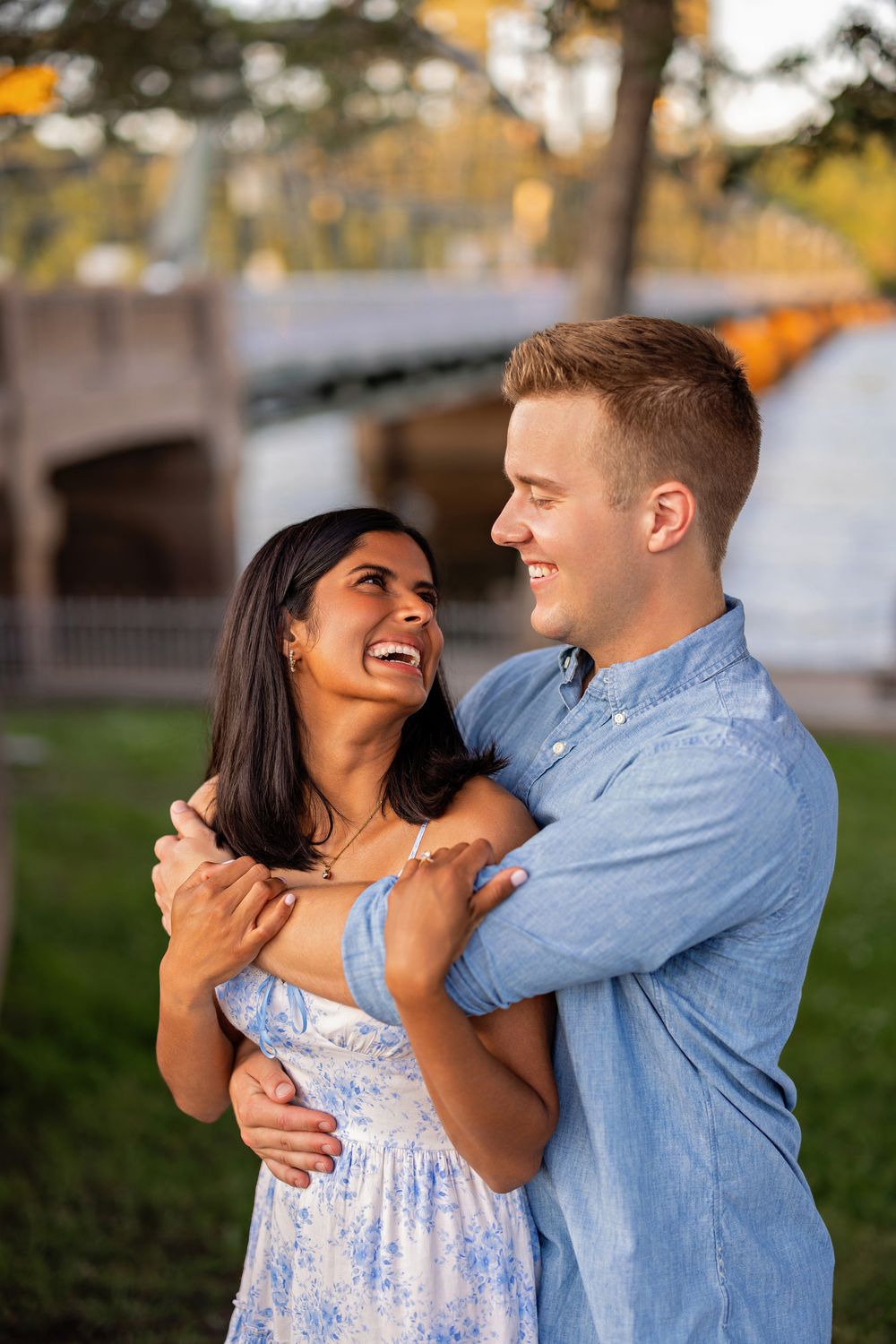 Laughing couple embracing face-to-face outdoors in golden light near a bridge, sharing a joyful moment.