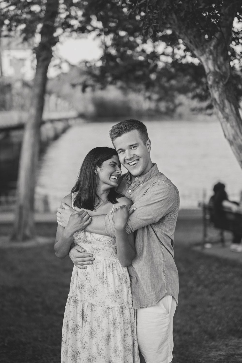 Black and white photo of a happy couple embracing and smiling in a park near a river.