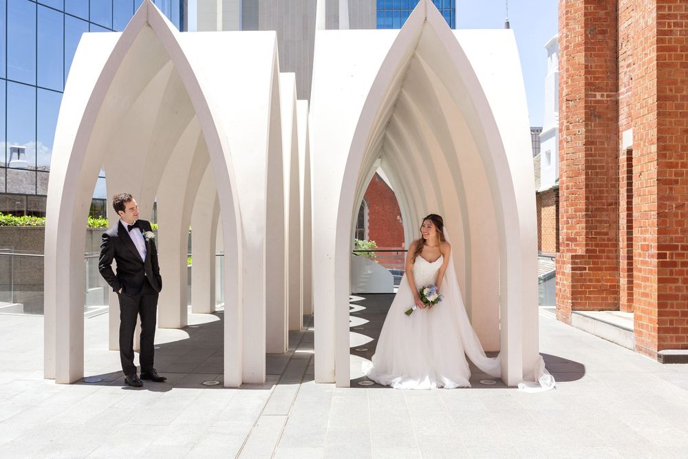 Bride and groom pose separately under elegant white Gothic arches outdoors on their wedding day.