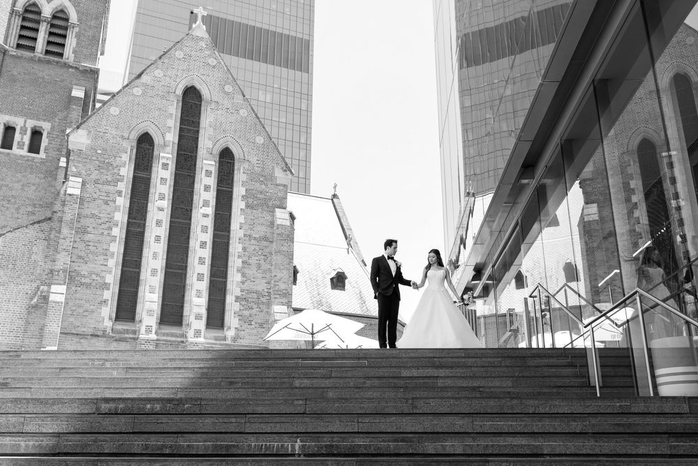 Black and white wedding photo of bride and groom holding hands on steps with church and skyscrapers in background.