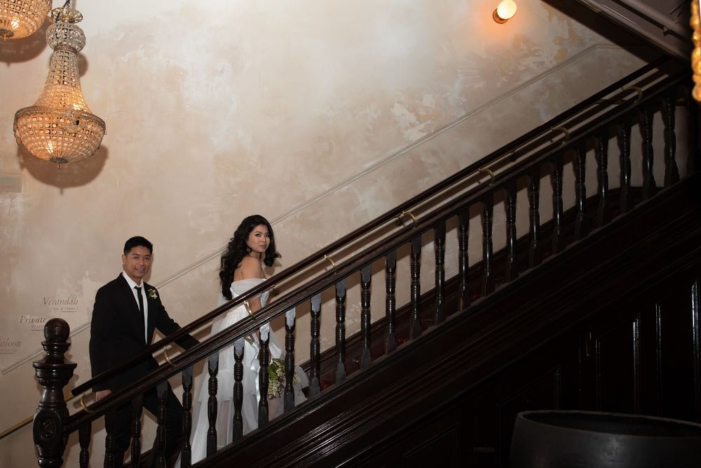 Bride and groom descend a grand wooden staircase under crystal chandeliers at wedding venue.