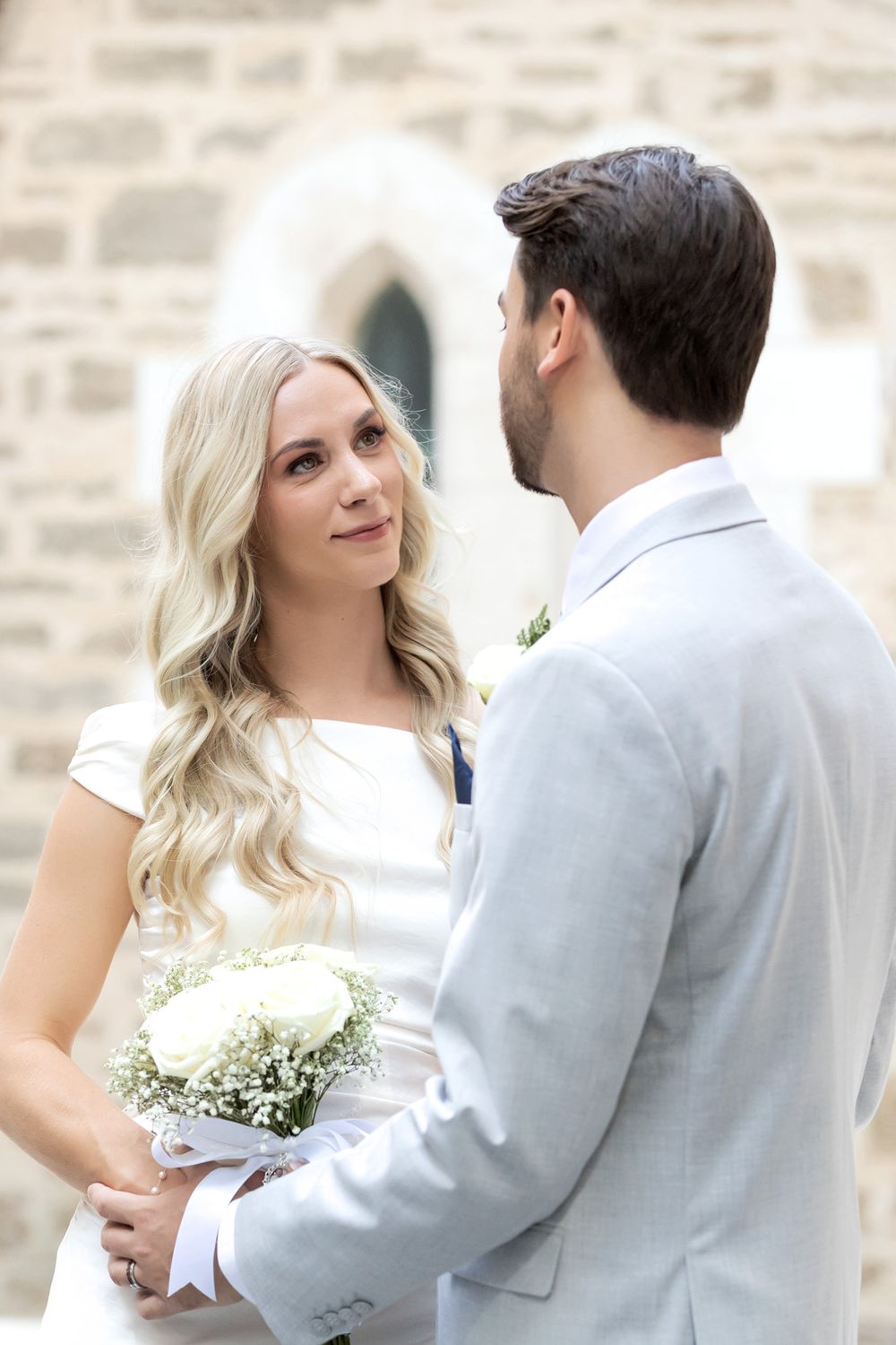Bride and groom exchanging vows outdoors, bride holding white rose bouquet, wearing wedding dress, groom in light gray suit.
