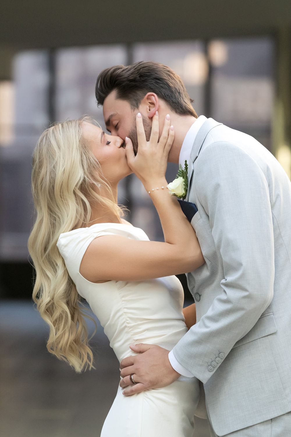 Bride and groom sharing a romantic kiss, bride in white dress with flowing blonde hair, groom in light gray suit with boutonniere.