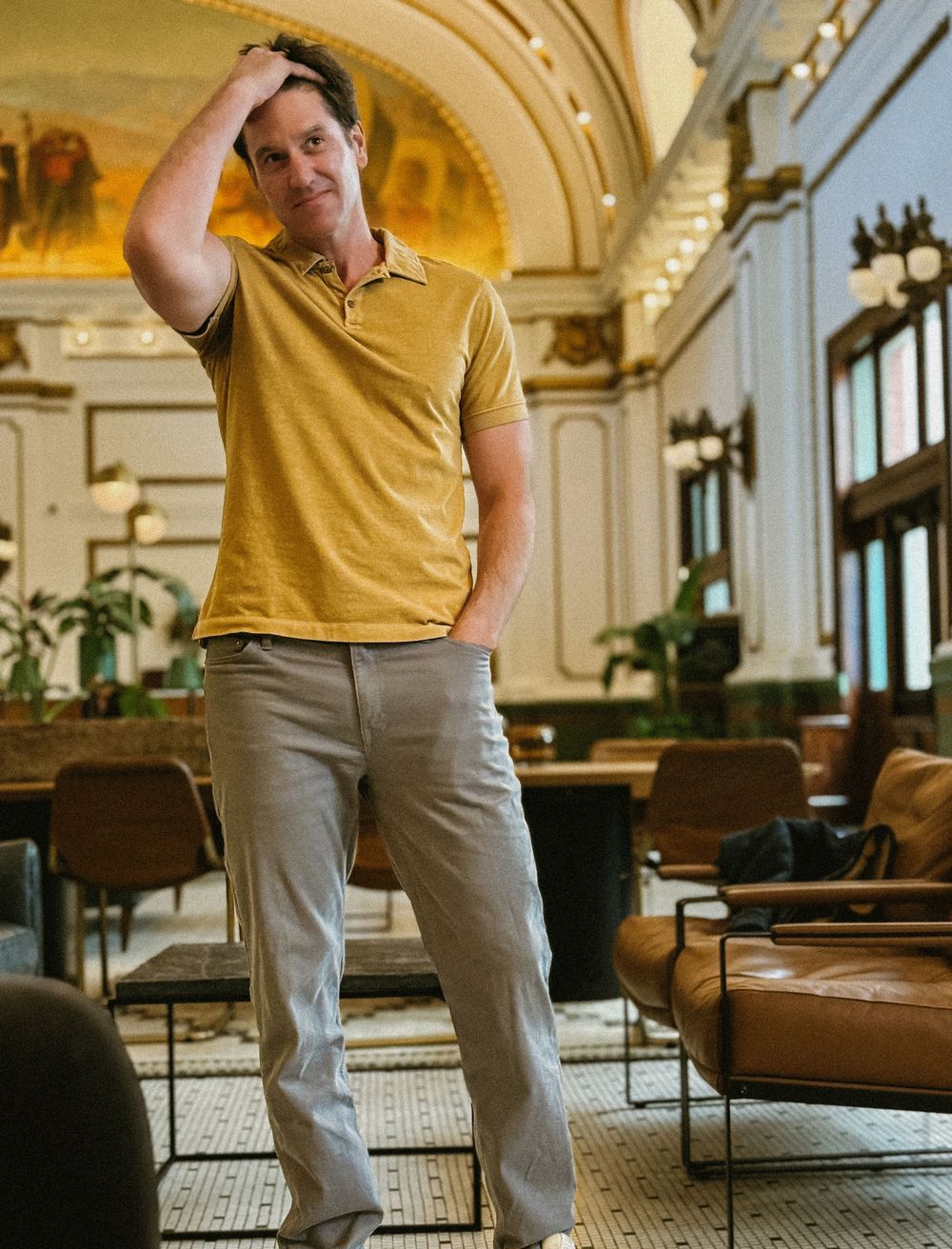 Man in mustard polo shirt running hand through hair posing in elegant hotel lobby interior.