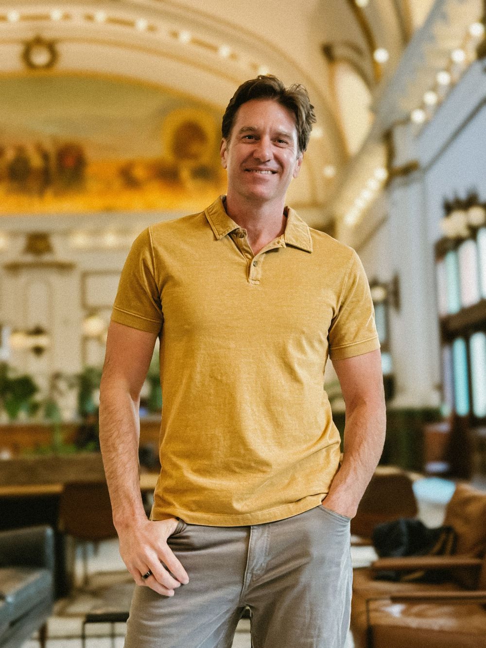 Smiling man in yellow polo shirt standing casually in grand ornate hotel lobby space.