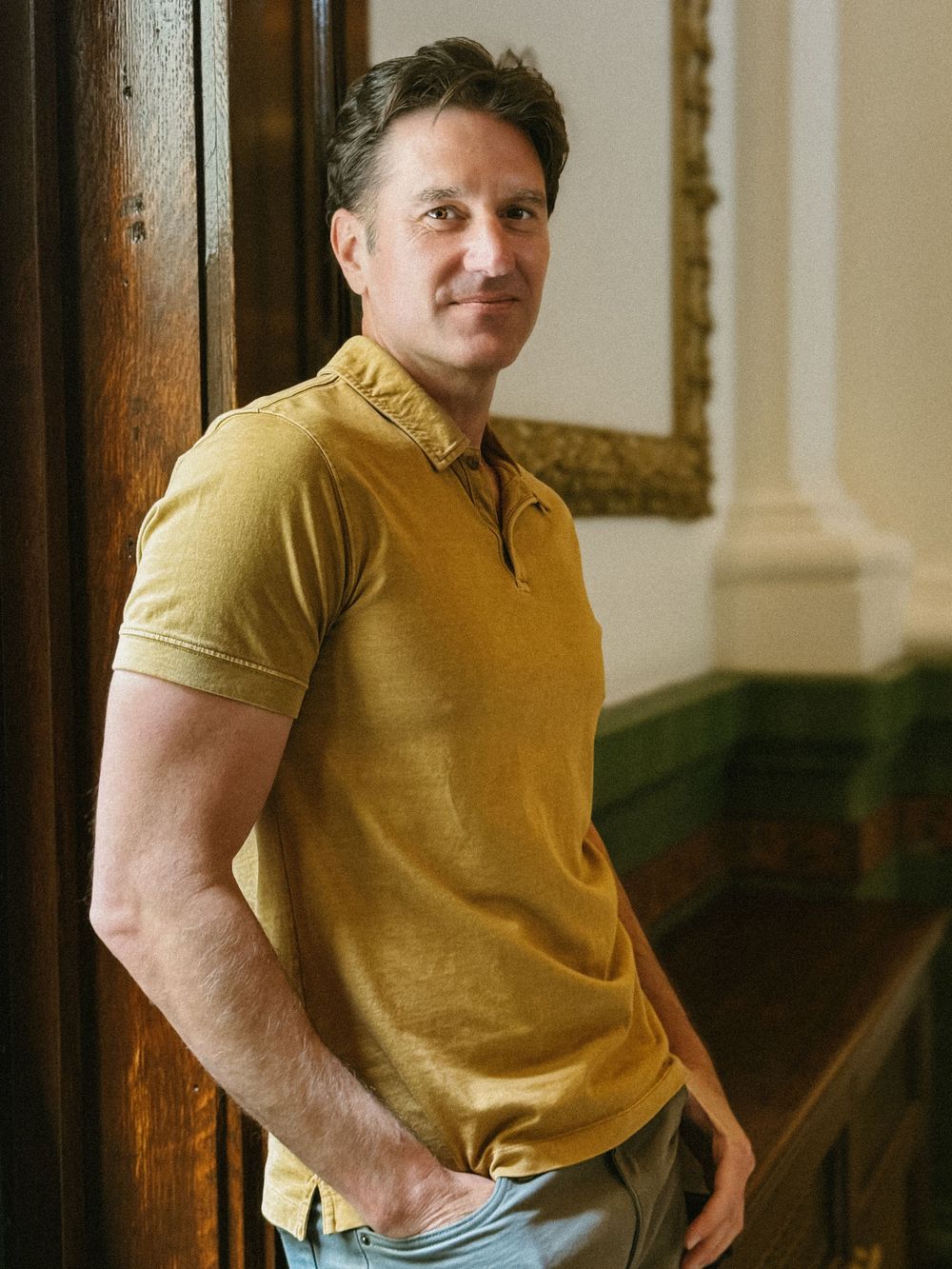 Man in mustard yellow polo shirt leaning against wooden wall beside gold-framed artwork.