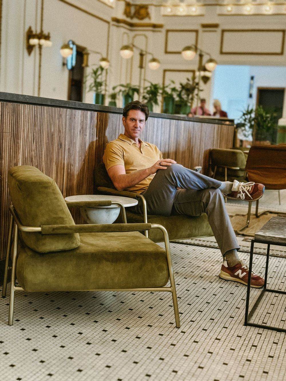 Man in yellow polo shirt relaxing in olive velvet chair in stylish hotel lobby.