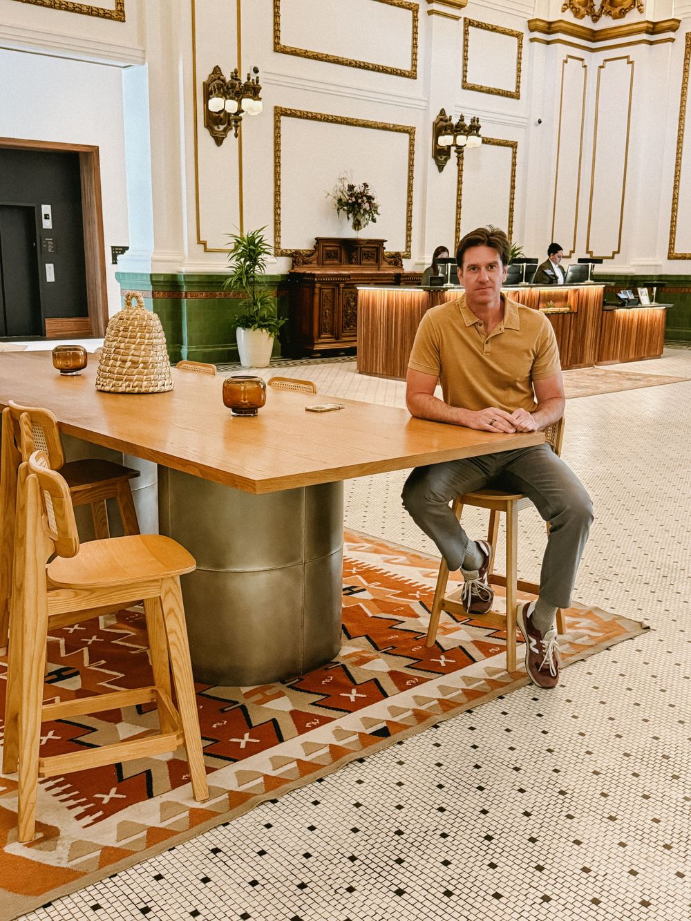 Man sits at wooden table with brass base in elegant hotel lobby with ornate white walls and patterned rug.