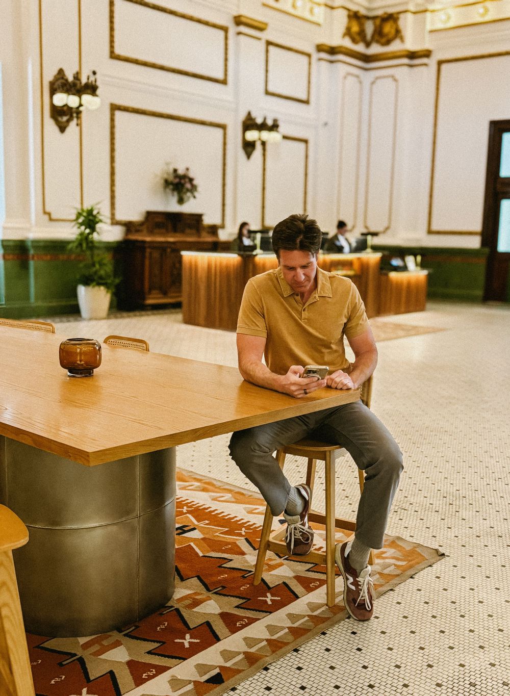 Man sits on stool using phone at wooden table in elegant hotel lobby with ornate decorations.