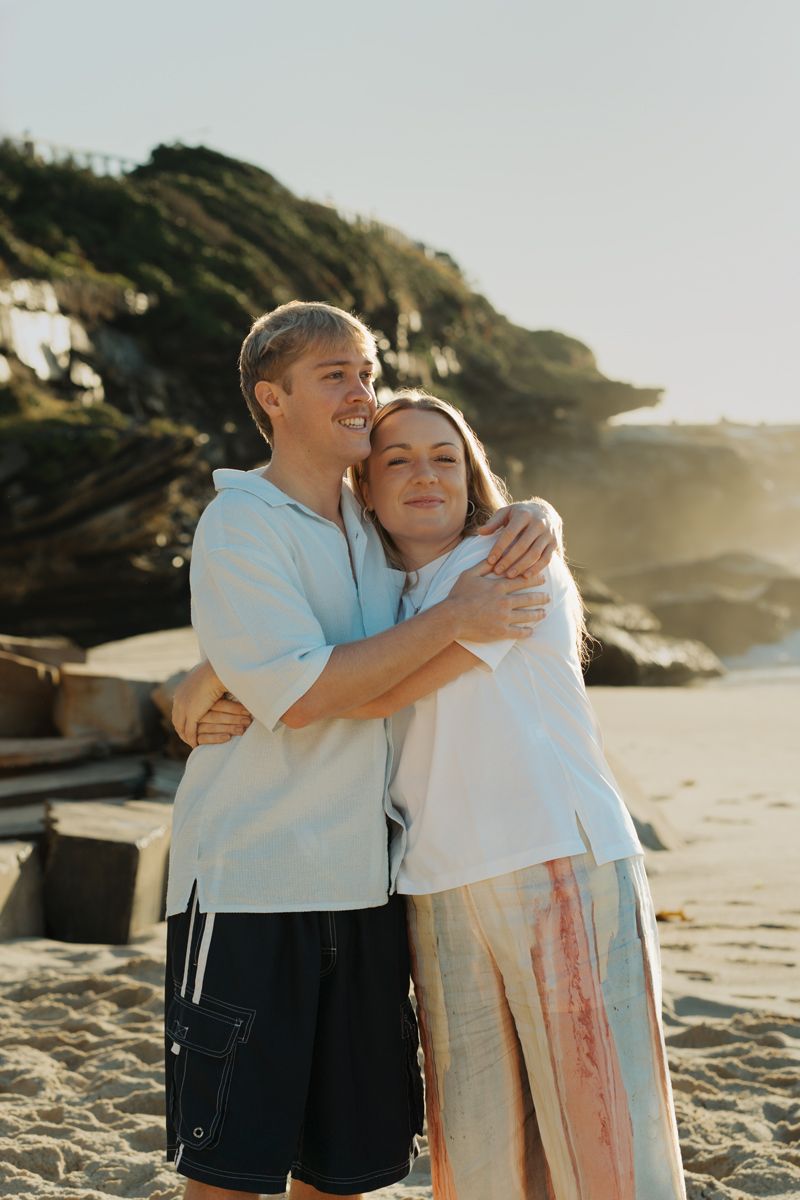 Brother and sister on a sunny beach with rocky cliffs, smiling warmly in casual summer outfits during a family photoshoot at Bronte Beach