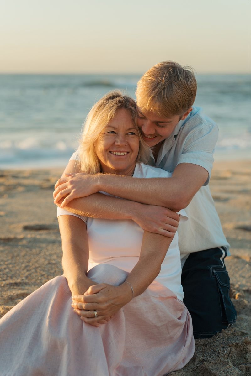 A young man hugs his mom on a sandy beach at sunrise photoshoot, both sitting together near ocean waves at Bronte Beach Sydney
