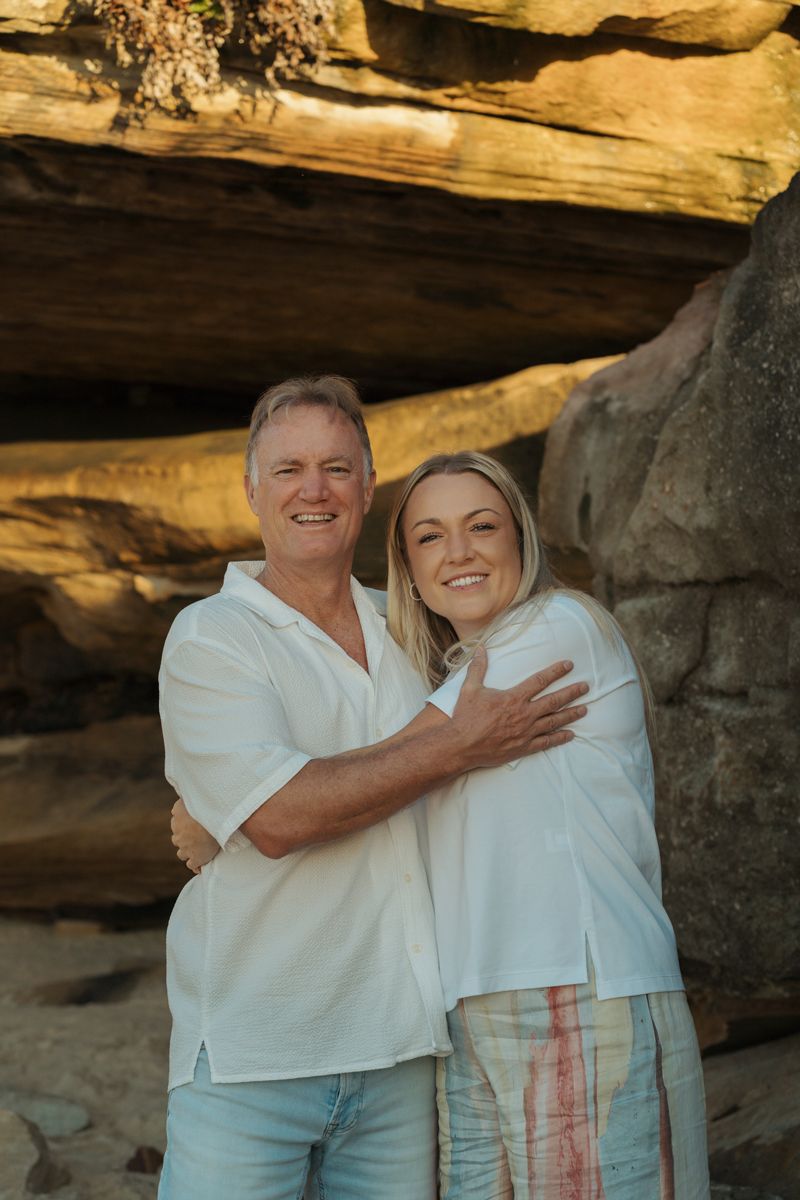Father and adult daughter hugging and smiling in front of large rock formation on Bronte beach in Sydney during a family photoshoot