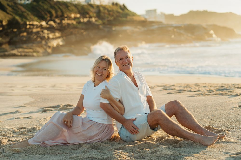 A smiling middle-aged couple sits together on a sandy beach at golden hour photoshoot wearing casual white tops at Bronte Beach Sydney