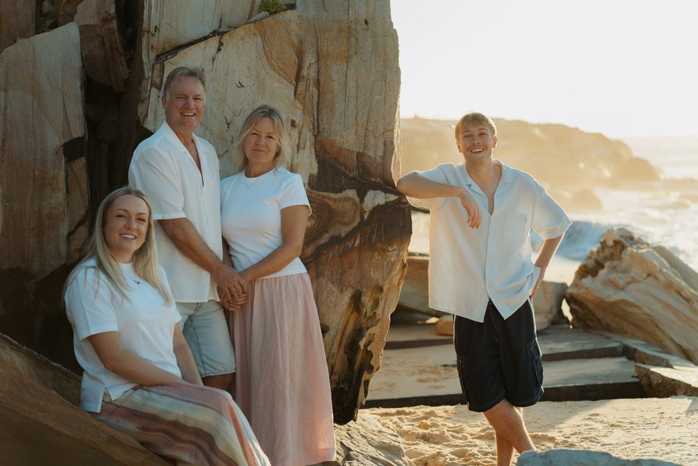 Family of four posing for family photoshoot at th beach at sunset, wearing white tops, with ocean waves in background in Bronte Beach Sydney