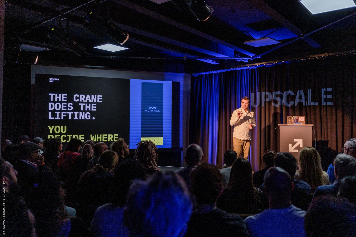 Speaker presenting on stage at Upscale event with projected text reading The Crane Does The Lifting. You Decide Where.