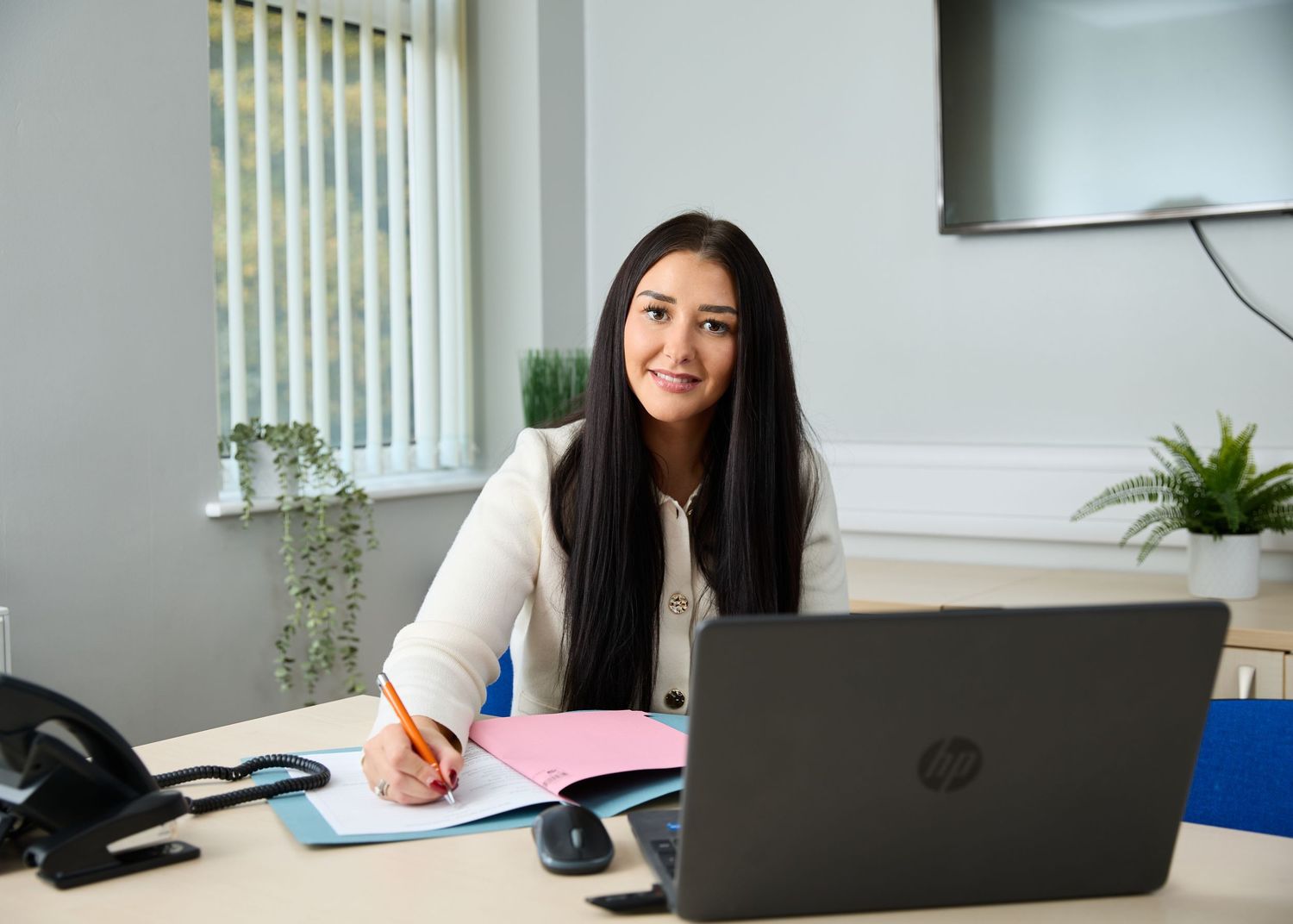 Young professional woman smiling at desk with HP laptop, notebook, and pen in a modern bright office.