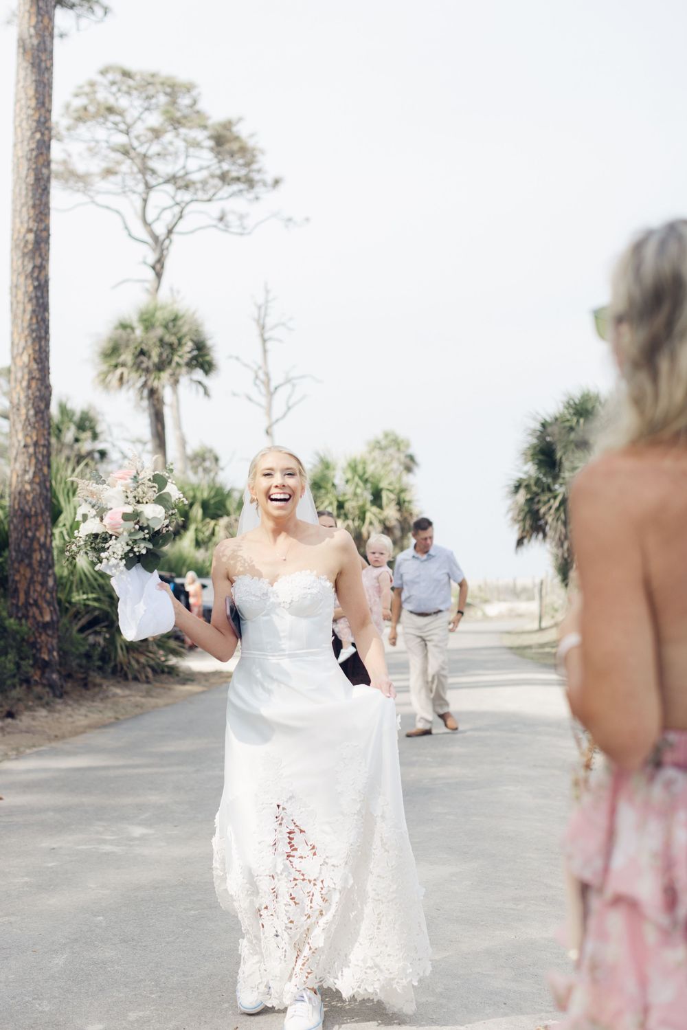 Laughing bride in white lace gown holding bouquet walks outdoors with palm trees in background on wedding day.