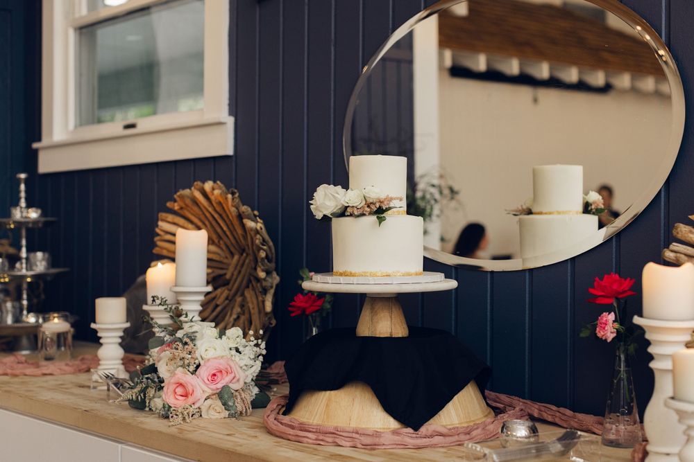 Elegant wedding dessert table with white tiered cake on gold stand, candles, floral arrangements, and round mirror against navy wall.