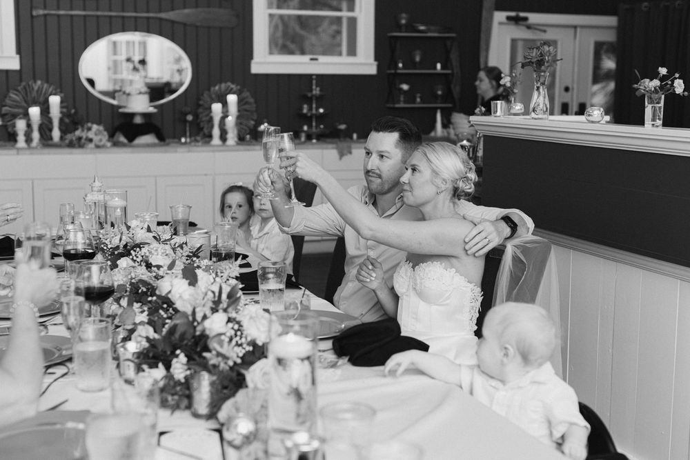 Black and white photo of a bride and groom toasting champagne at their wedding reception dinner table.