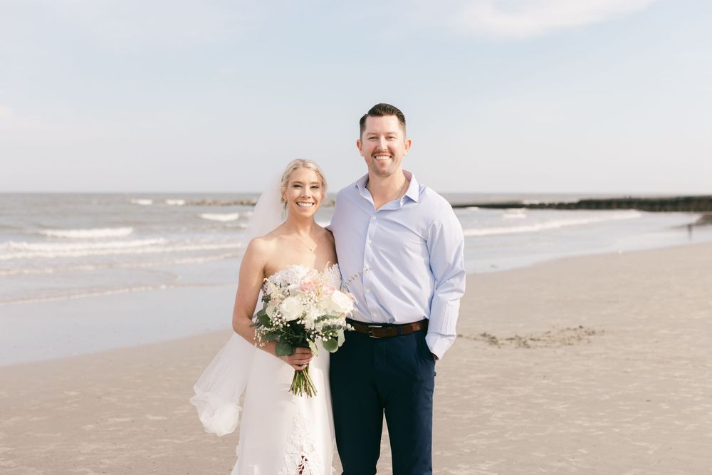 Bride and groom smiling together on a sandy beach, bride holding a white floral bouquet with ocean waves in the background.