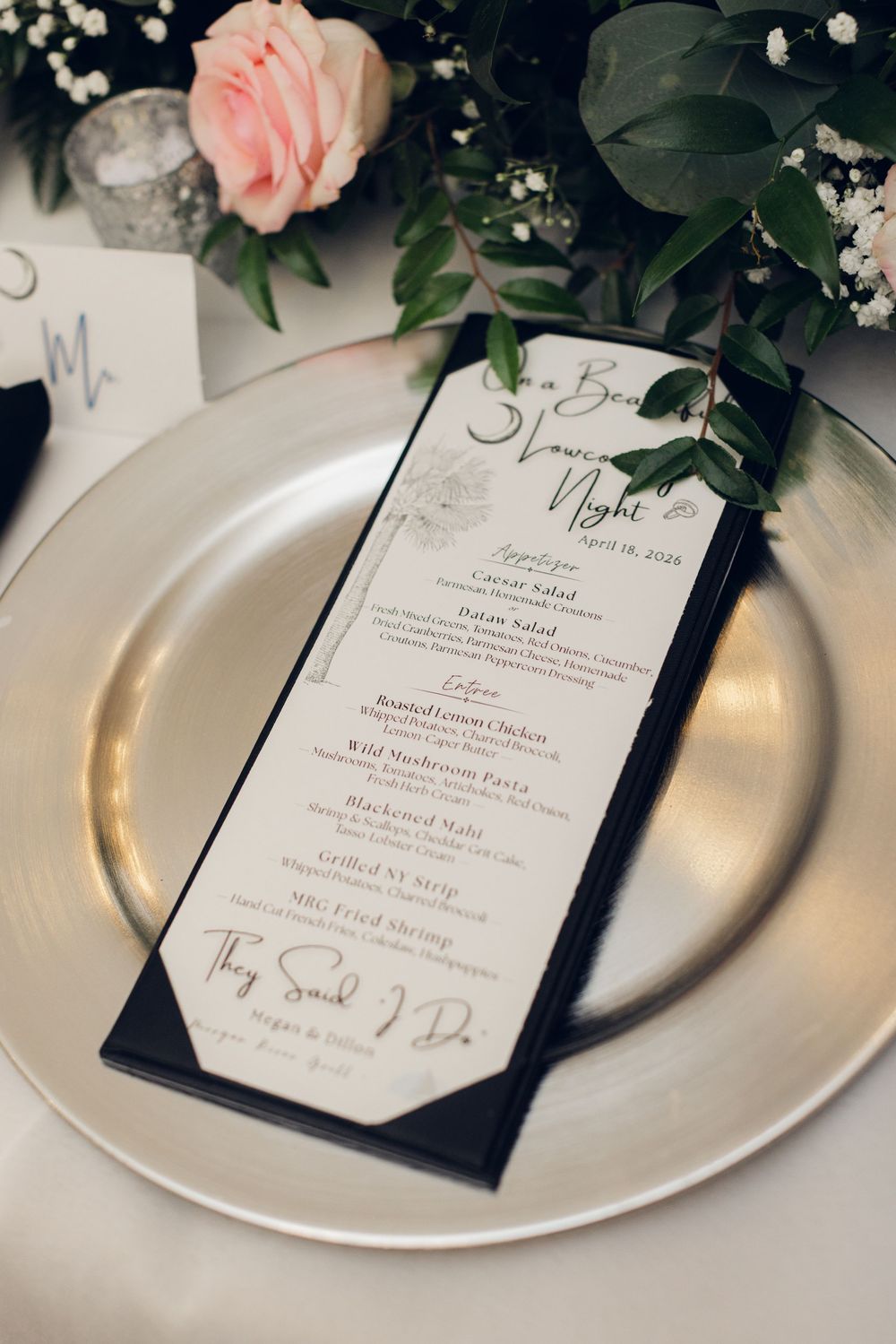 Elegant wedding dinner menu on silver charger plate with pink roses and greenery centerpiece.