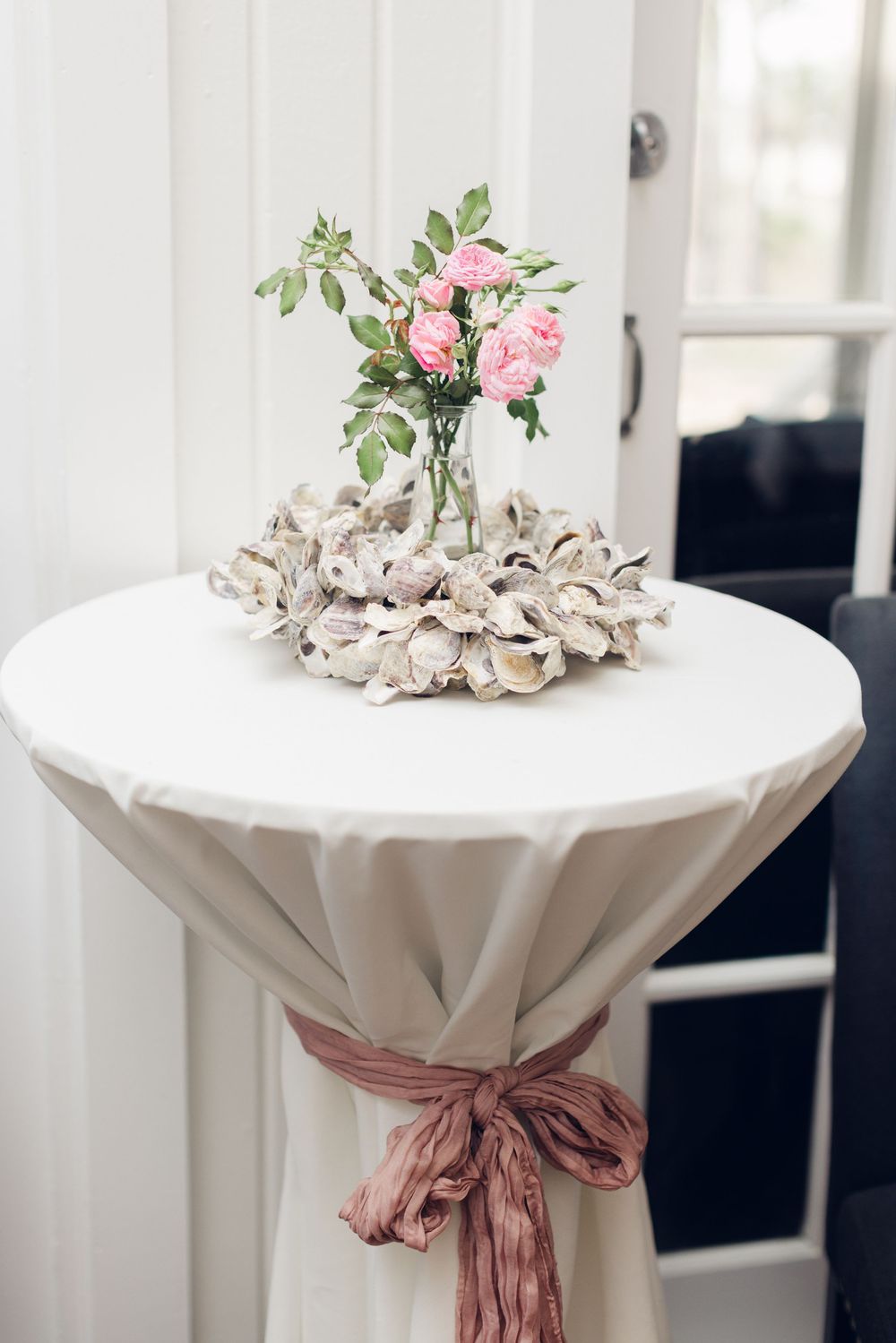 Elegant cocktail table with white linen, dusty rose bow, oyster shell wreath, and pink roses in glass vase.
