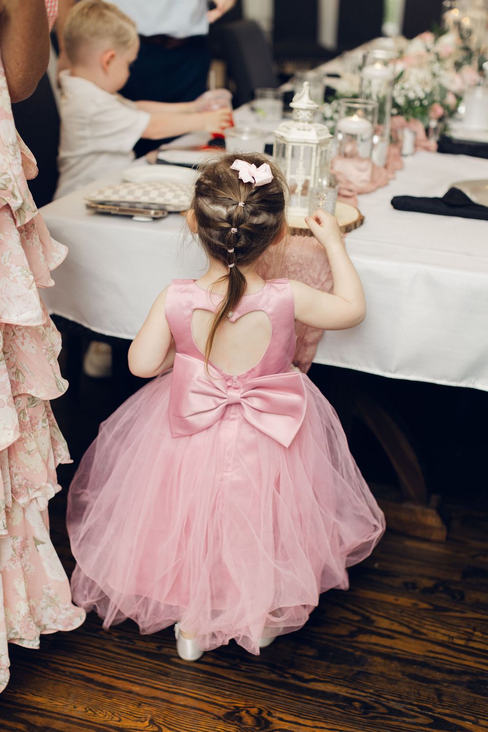 A little girl in a pink tulle dress with a bow back stands at a decorated wedding reception table.