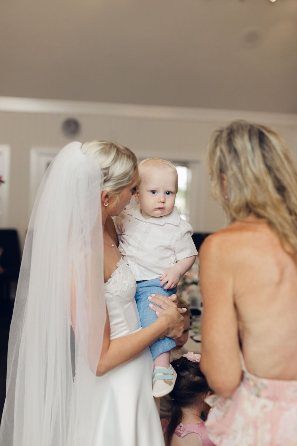 Bride in white gown and veil holds a baby in white outfit while chatting with a woman in a floral dress.