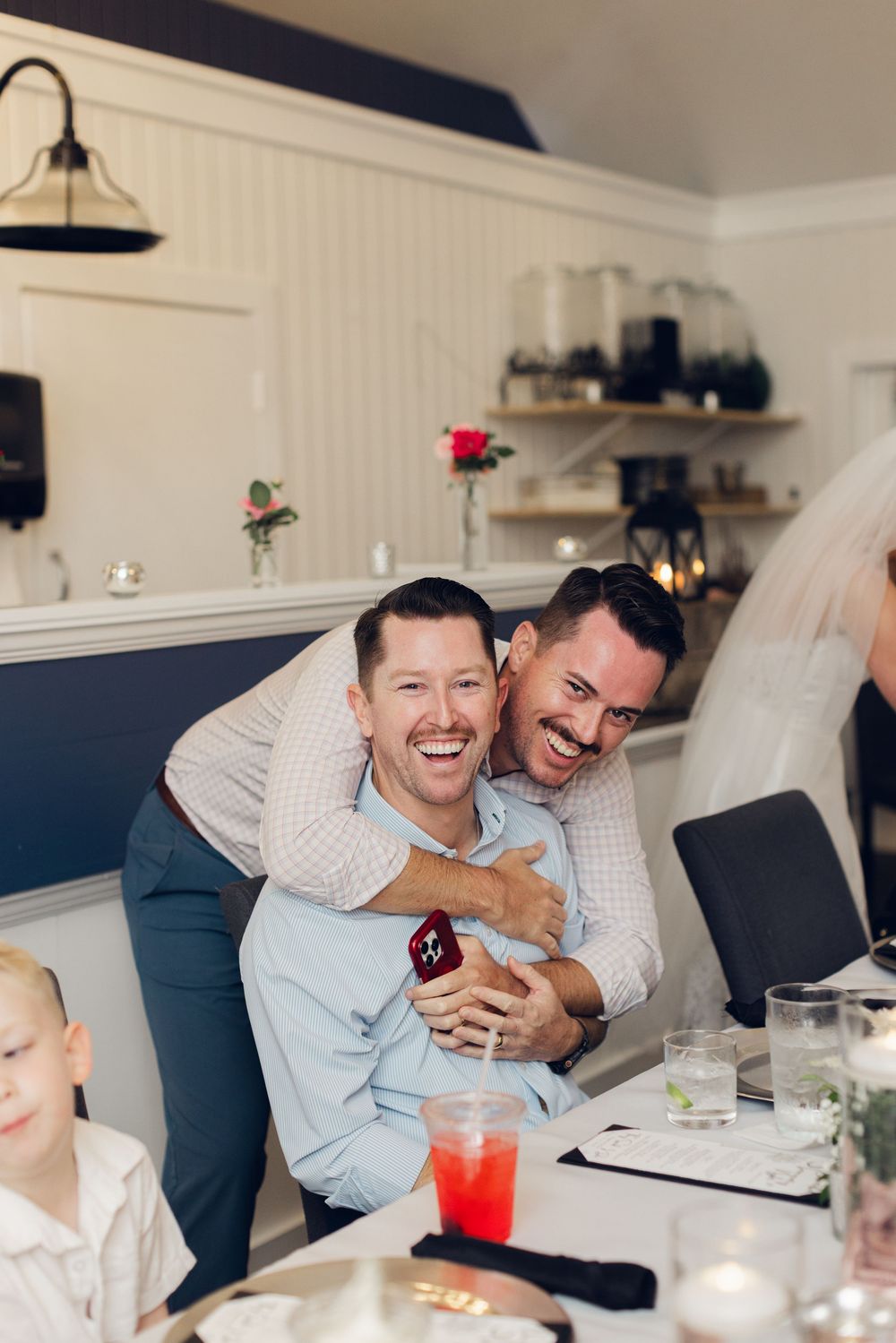 Two men laughing joyfully at a wedding reception table, one hugging the other from behind in a celebratory moment.