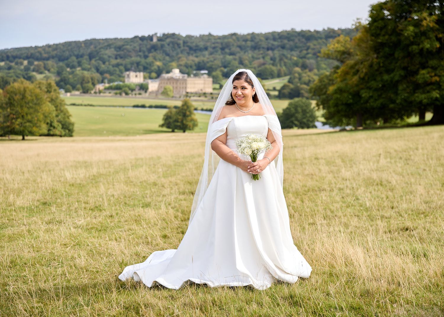 Bride and groom holding hands walking through a lush green meadow with a grand estate and rolling hills in the background.