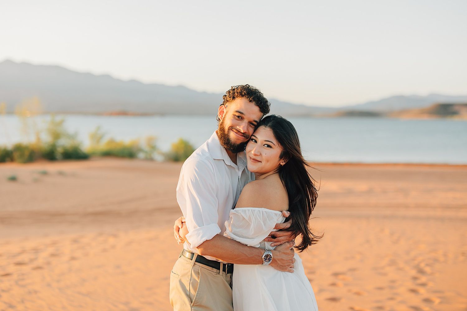 Happy couple hugging on golden sand beach at sunset with serene lake and mountains behind them.
