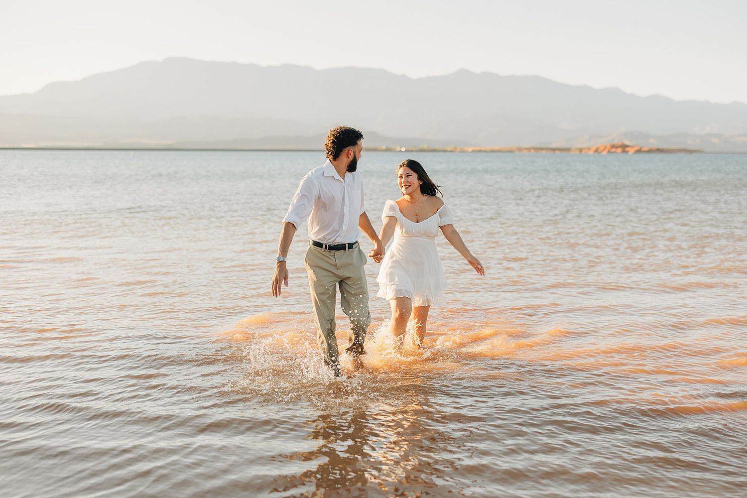Couple in white outfits walking hand-in-hand through shallow lake water with mountains in the background.