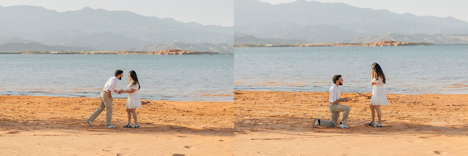 Couple on sandy beach with mountains, man kneeling proposing to woman in white dress near blue water.