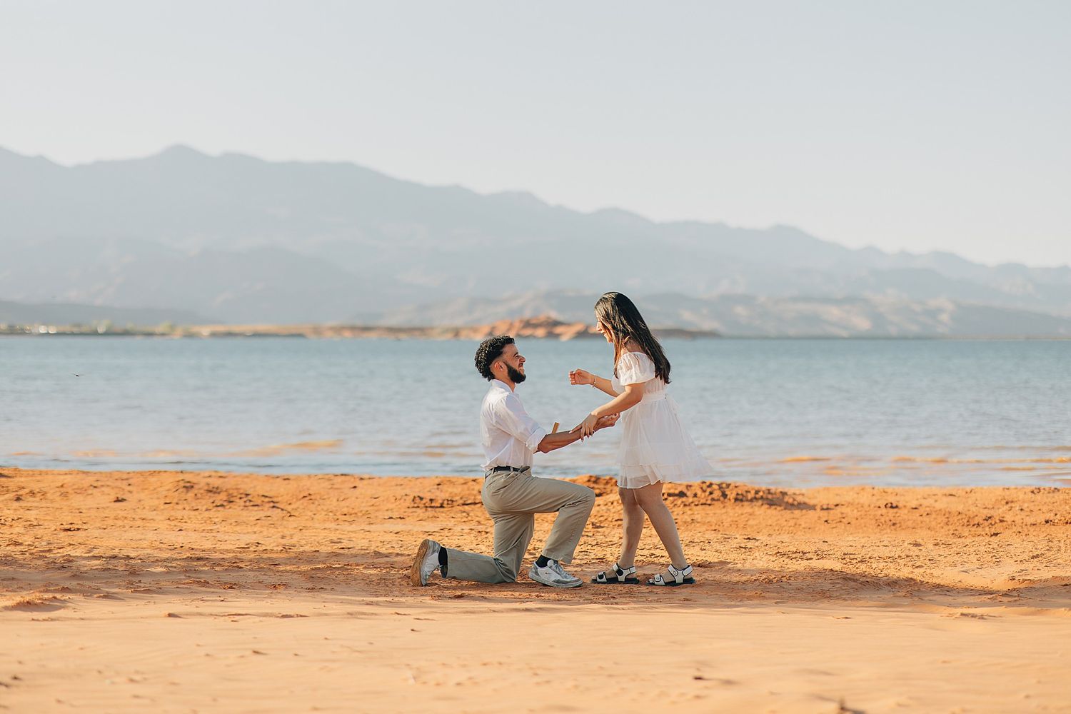 Man proposing to woman on sandy beach with mountains and lake in background on a sunny day.