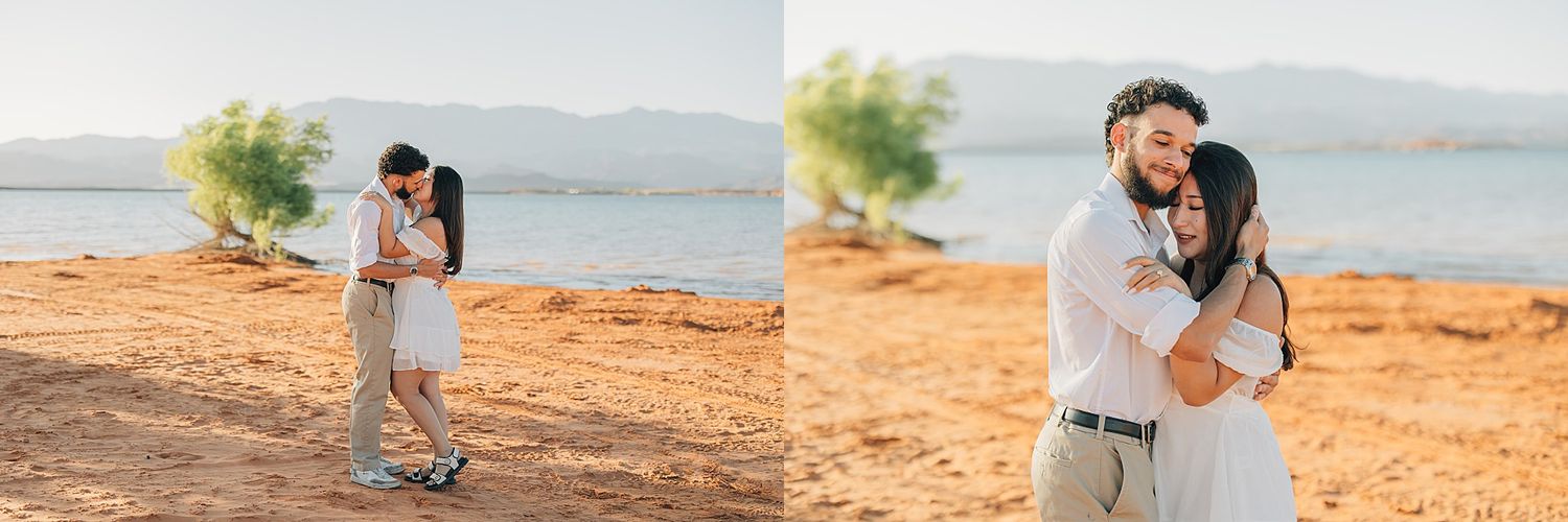 Couple embracing on sandy beach shore with trees and calm lake in background during golden hour.