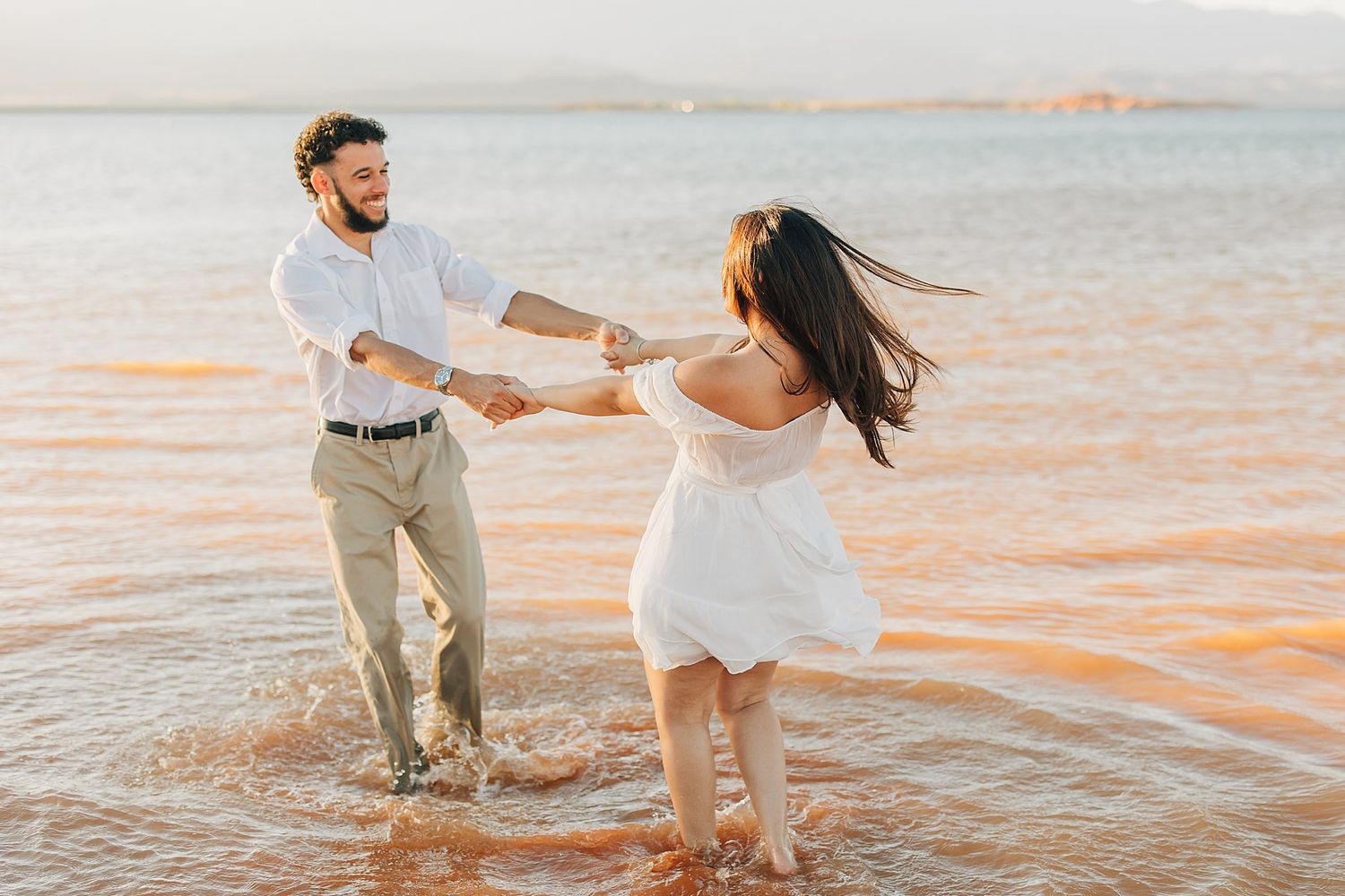 Couple dancing and splashing in ocean waves at sunset, woman in white dress, man in white shirt and khakis.