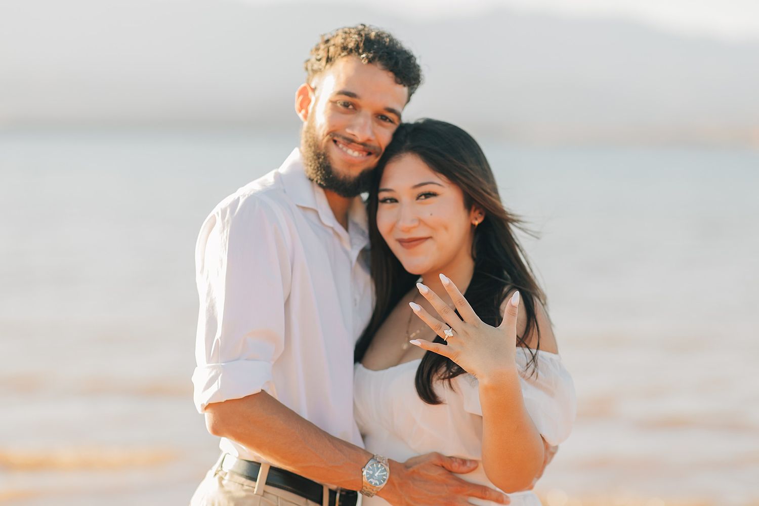 Happy engaged couple poses on beach as woman shows off her sparkling engagement ring.