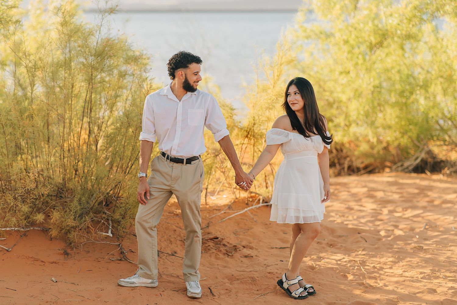 Couple holds hands walking on red sand near lake, woman in white dress, man in white shirt and khaki pants.