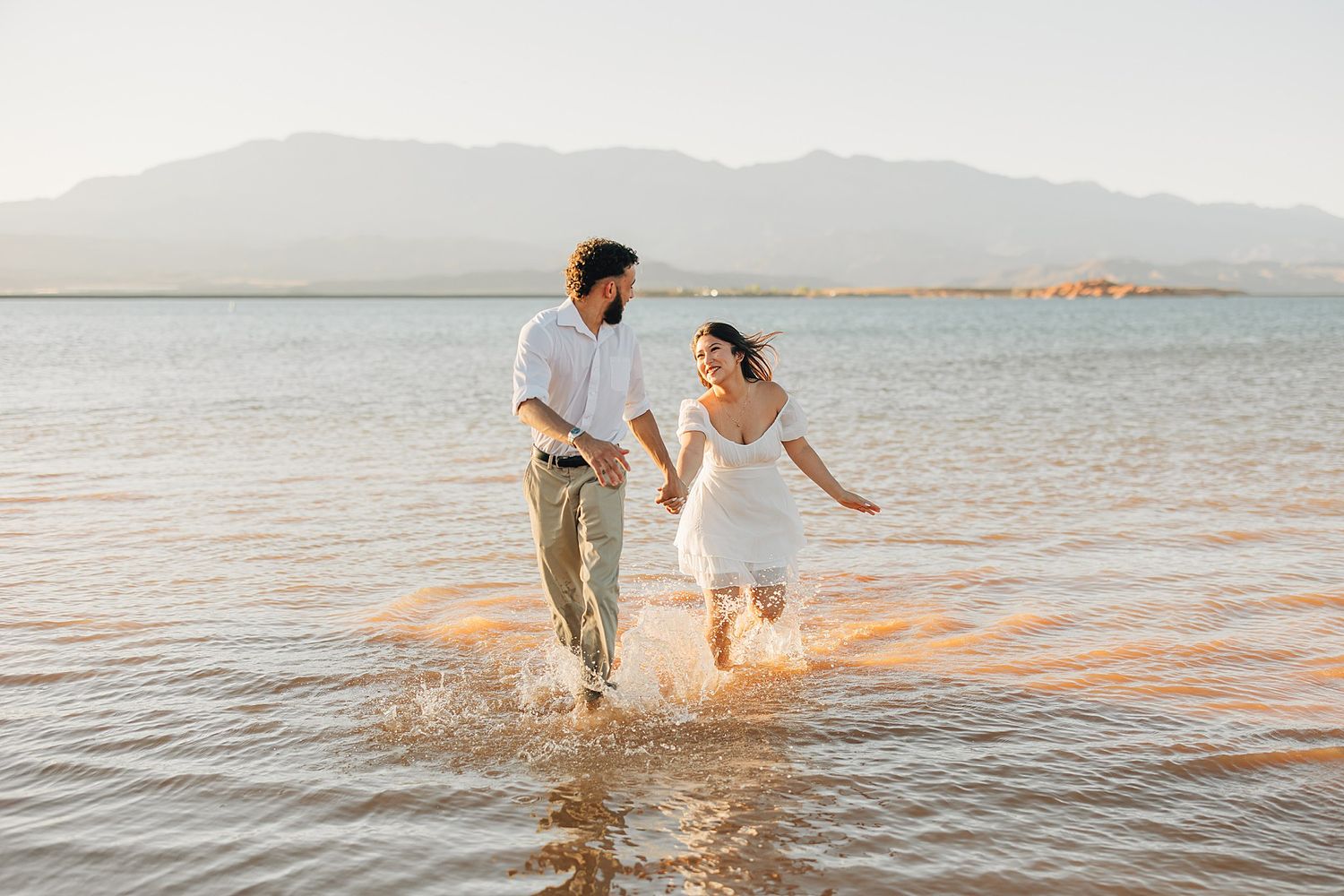 Couple holding hands running through shallow water at golden hour with mountains in the background.