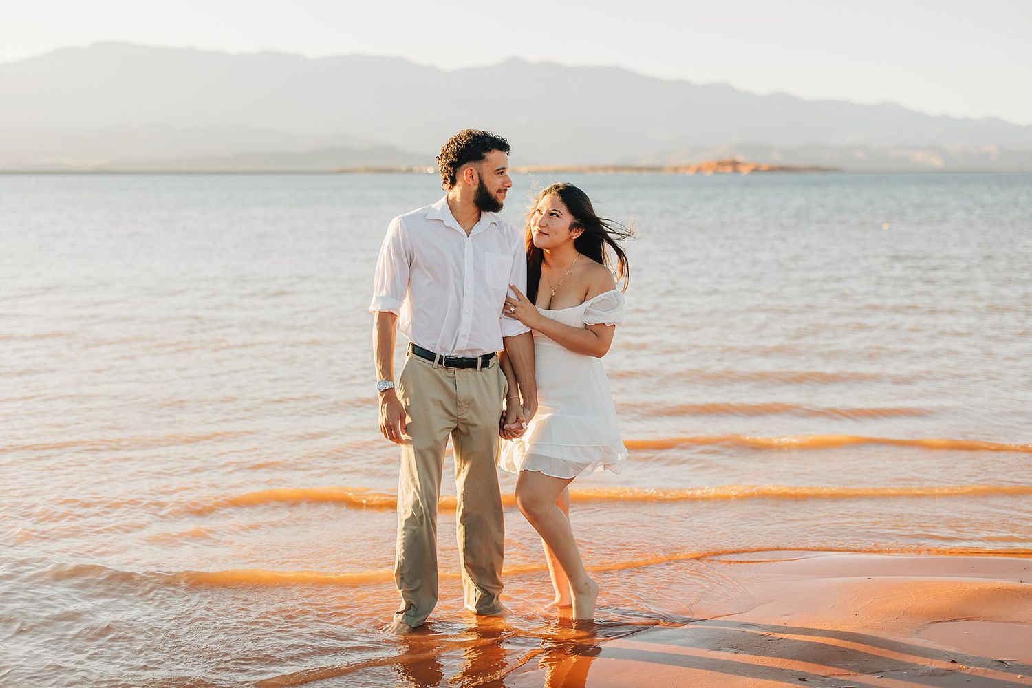 Couple standing in shallow water at golden hour, wearing white outfits, smiling at each other with mountains in background.