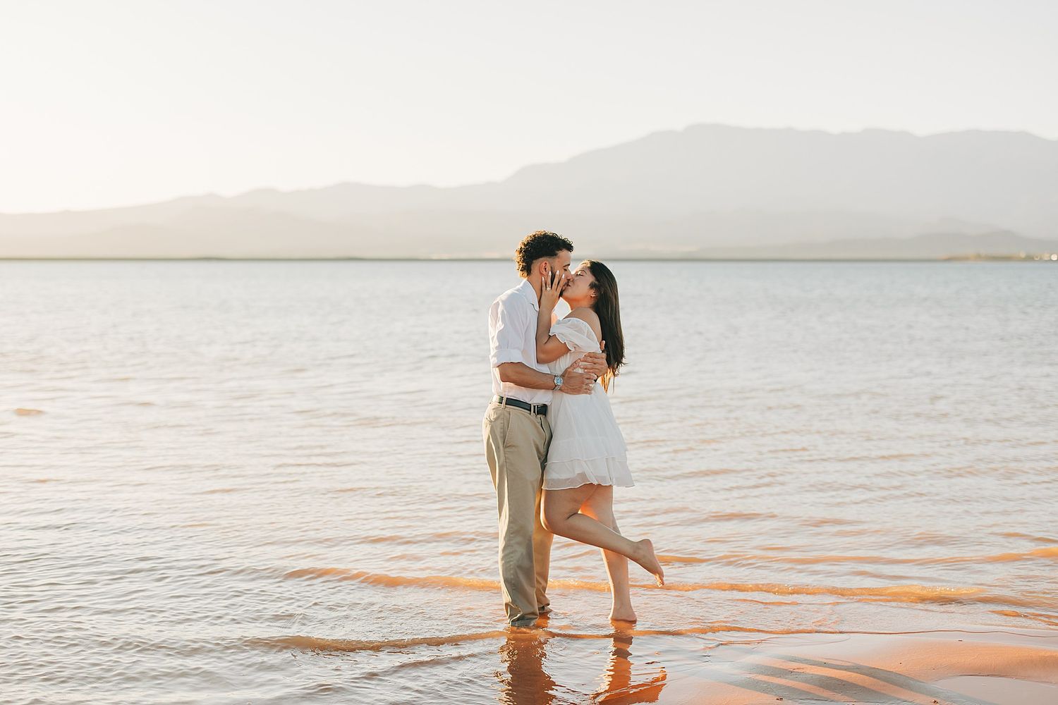 Couple sharing a romantic kiss while standing in shallow water at golden hour with mountains in the background.