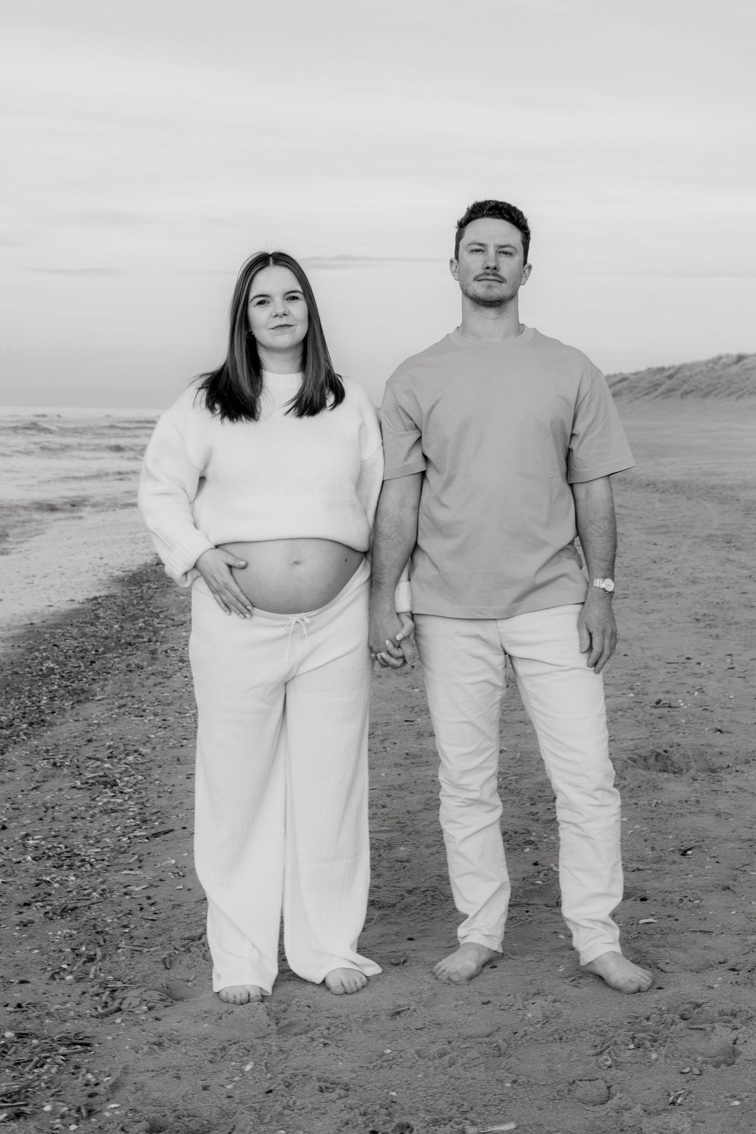 Black and white photo of a pregnant couple holding hands on a beach, woman cradling baby bump, both barefoot.