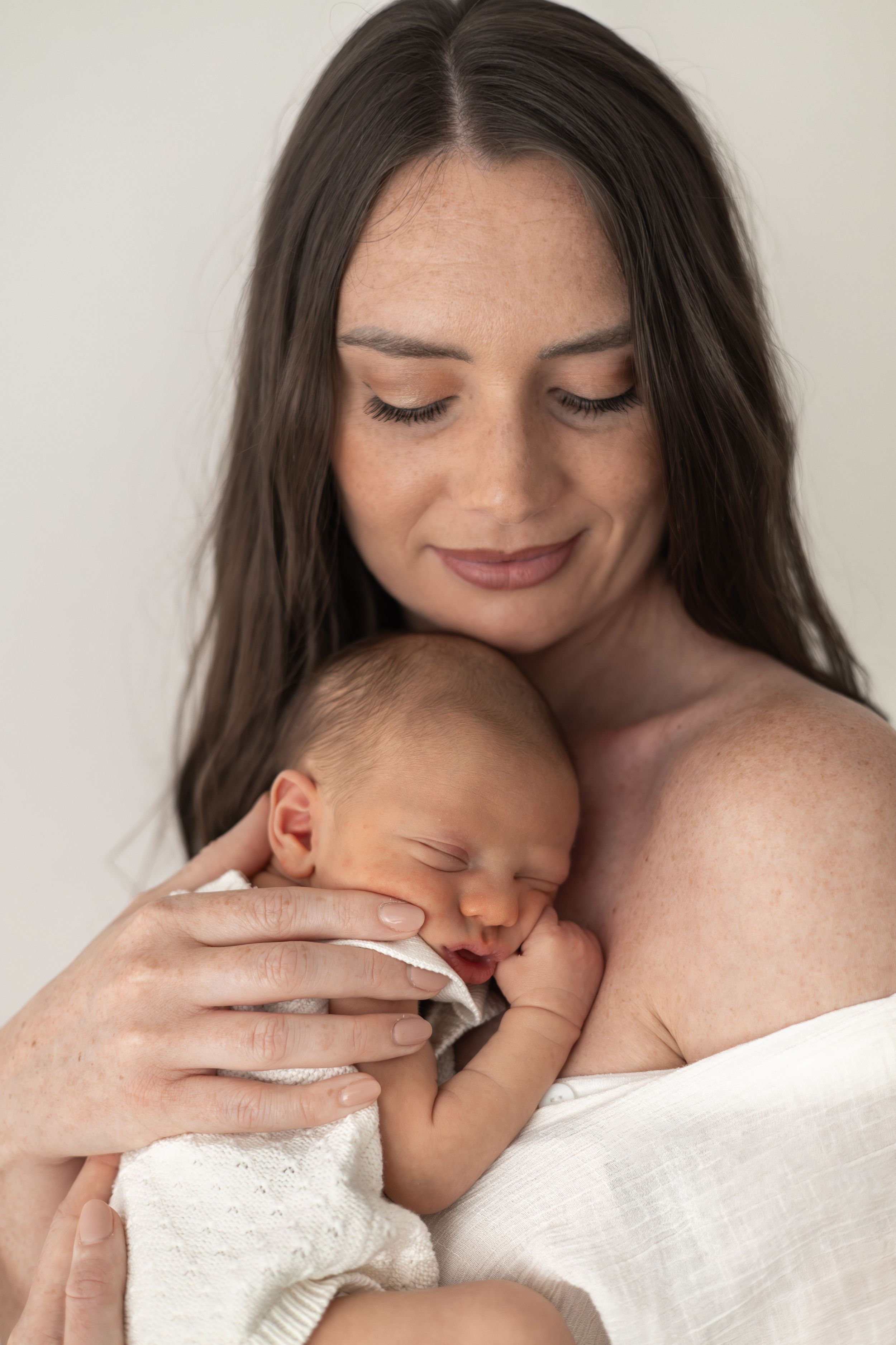 A smiling mother tenderly holds her sleeping newborn baby wrapped in a white blanket during newborn photographer Nottingham