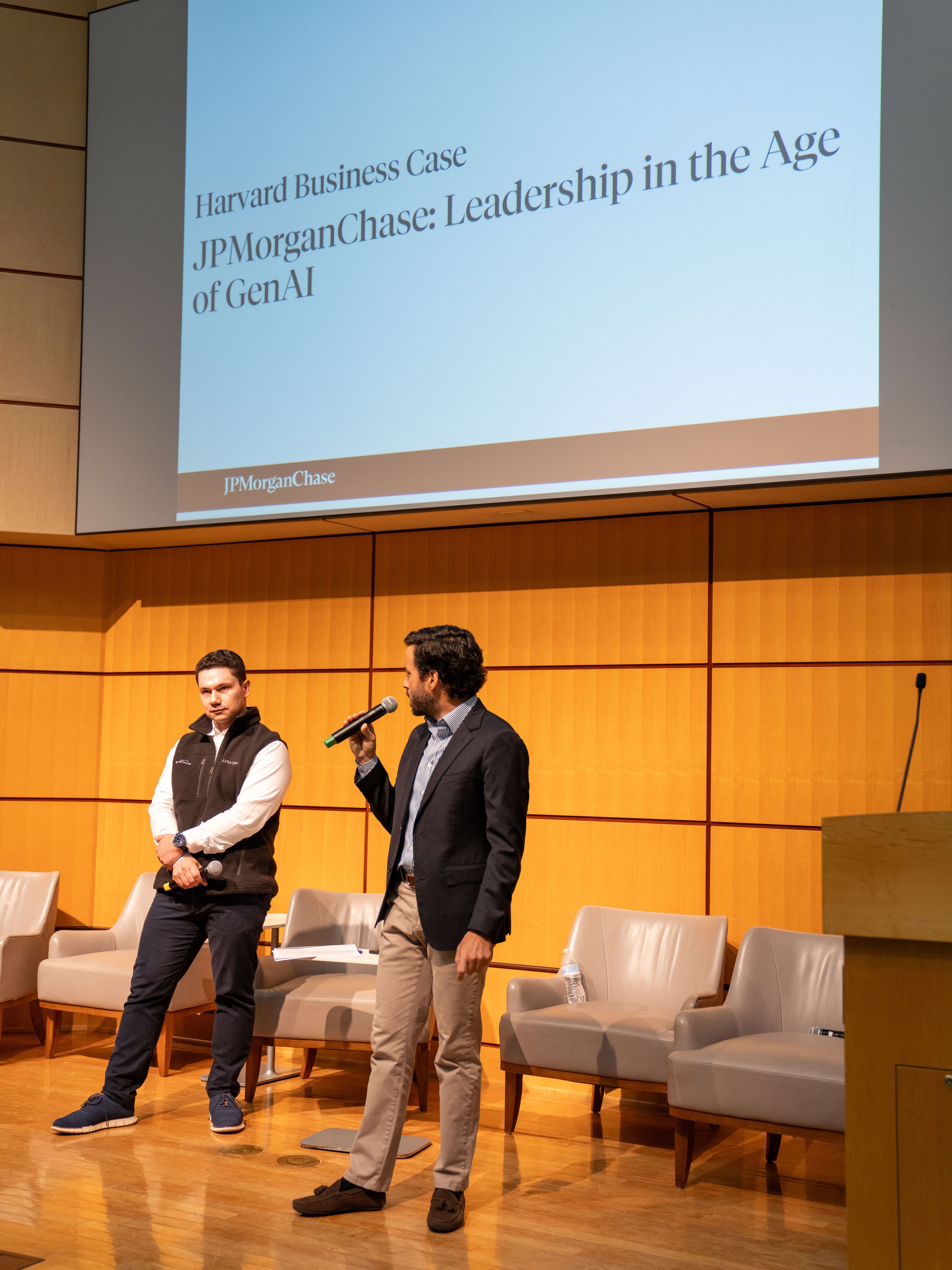 Two men present a Harvard Business Case on JPMorganChase Leadership in the Age of GenAI on a modern auditorium stage.