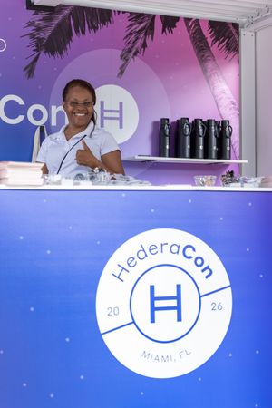 A smiling woman gives a thumbs up at the HederaCon 2026 Miami, FL registration booth with purple branding.