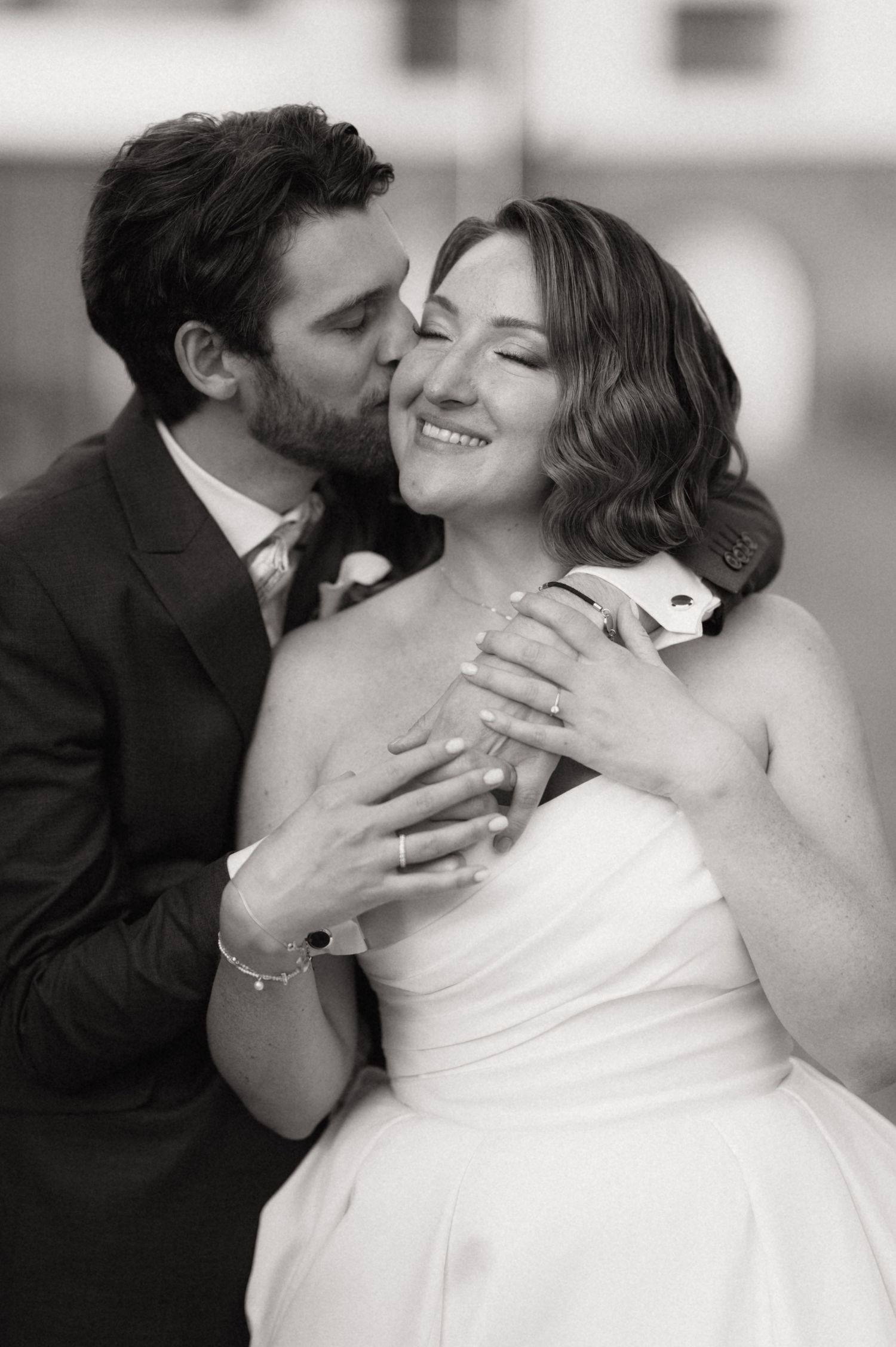 Black and white wedding photo of groom kissing bride on cheek while embracing her from behind, bride smiling with eyes closed.